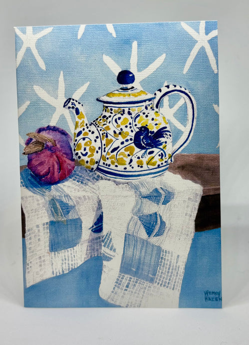 A yellow, white and blue teapot with a blue bird motif.  There is a red onion for color and interest as well as star fish images against a blue wall.