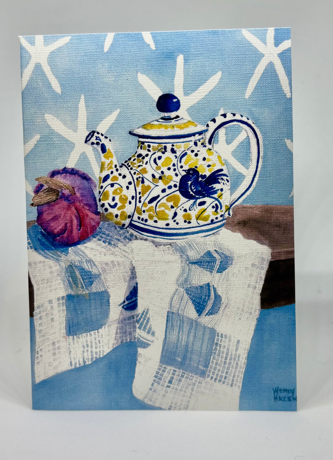 A yellow, white and blue teapot with a blue bird motif.  There is a red onion for color and interest as well as star fish images against a blue wall.