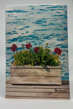 Load image into Gallery viewer, Lake water in the background and sitting on this weathered dock is a lovely weathered flower box with brilliant red geraniums.
