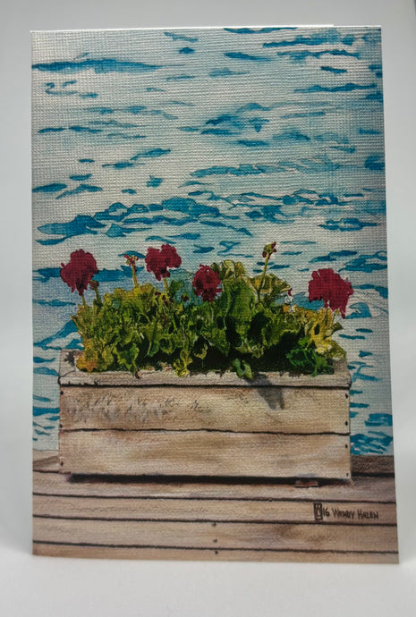Lake water in the background and sitting on this weathered dock is a lovely weathered flower box with brilliant red geraniums.