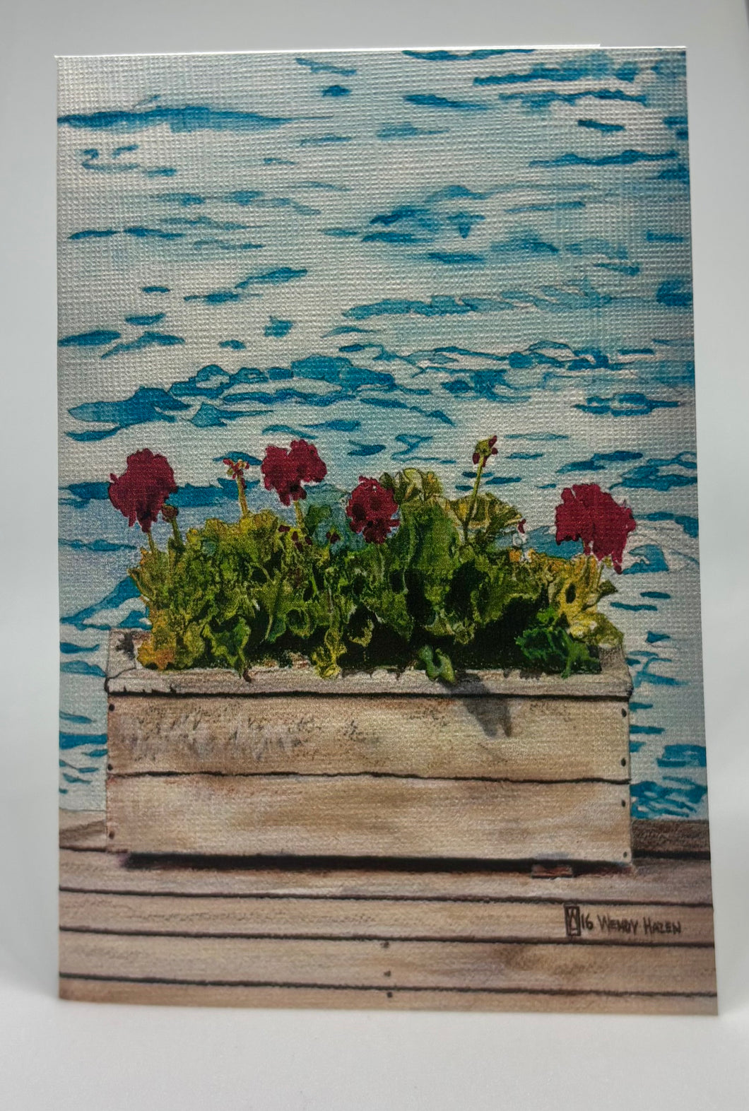 Lake water in the background and sitting on this weathered dock is a lovely weathered flower box with brilliant red geraniums.