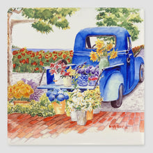 Load image into Gallery viewer, A ceramic tile of a blue truck with flowers on brick walkways and cobblestone street background with a view to the sea.
